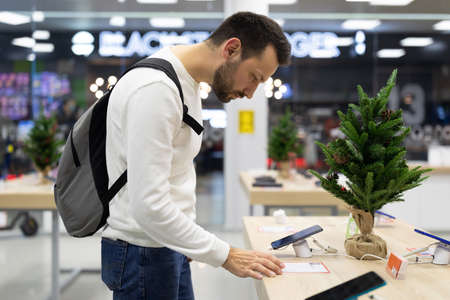 Photo of an adult handsome doubtful thinking brunette man with stubble in a white sweatshirt chooses a mobile phone in an electronic store in a shopping mall. Shop conceptの写真素材