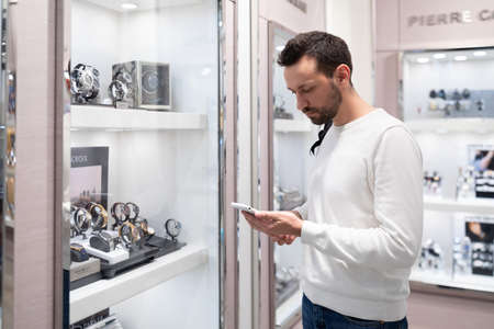 Minsk, Belarus - Nov 27, 2021: Photo of an adult handsome brunette man in a white sweatshirt and with a backpack chooses a stylish wrist watch in a store in a shopping center and using mobile phoneのeditorial素材