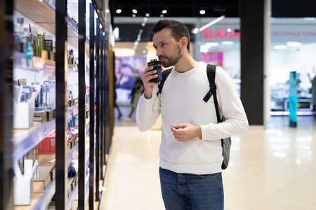 Minsk, Belarus - Nov 27, 2021: Photo of an adult handsome brunette man with stubble in a white sweatshirt chooses perfumery and adekolon in a perfume store in a shopping centerのeditorial素材
