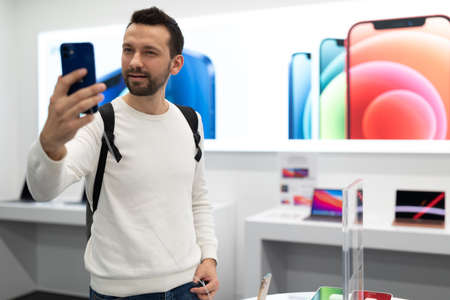 Minsk, Belarus - Nov 27, 2021: Photo of young handsome brunet man with stubble in a white sweatshirt chooses a mobile phone in an electronic store in a shopping mall. Smartphone shop conceptのeditorial素材