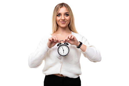 Photo of a beautiful pretty positive young blonde woman with makeup in a stylish white knitted sweater and black pants showing alarm clock isolated on a white background with empty space for textの写真素材