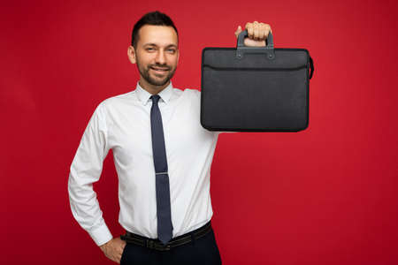 Photo of handsome brunet businessman holding his briefcase and smiling while wearing a white shirt...