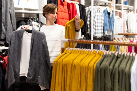 Photo of young attractive brunette woman with a short haircut in a white sweater chooses stylish and...