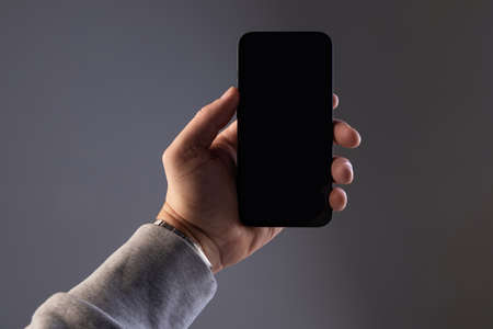 a male hand in a gray sweatshirt holds a black rectangular modern smartphone on a gray isolated background, a screen with space for copy-spaceの写真素材