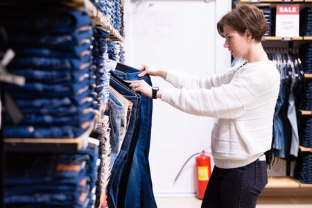 Photo of young attractive brunette woman with a short haircut in a white sweater chooses stylish and casual jeans and denim clothes in a store in a shopping centreの写真素材