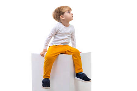 little child boy blond sits on a white cube and looks up to the right on a white isolated backgroundの写真素材