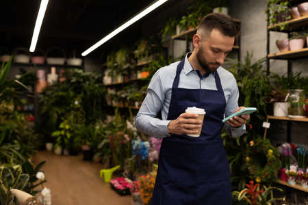 an employee in a flower shop with a glass of coffee watches the news on his phoneの写真素材