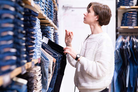 Photo of young brunette woman with a short haircut in a white sweater chooses stylish and casual jeans and denim clothes in a store. Shopping conceptの写真素材