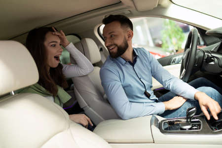 a man and a woman in a car dealership are sitting inside the car, being surprised by new...