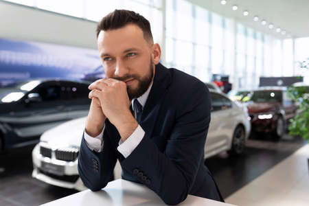 successful businessman in a car dealership next to new cars looks thoughtfully into the distanceの写真素材