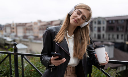 young blonde woman to mobile phone hands and a glass of coffee listens to music in headphones closing her eyes with pleasure on the background of the urban landscapeの写真素材