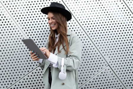 young cheerful woman in a black hat with headphones and a tablet smiling against the background of an aluminum facade Yes of an office buildingの写真素材