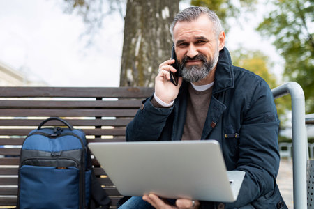 an elderly business man speaks on a mobile phone while working on a laptop online in a park on a...
