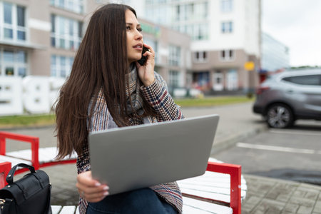 a young woman on the way to the office works on a laptop and makes a phone call on the background of the cityの写真素材