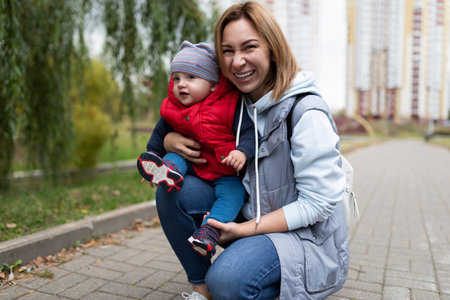 portrait of a young mother woman with her one-year-old child in her arms against the backdrop of a city parkの写真素材