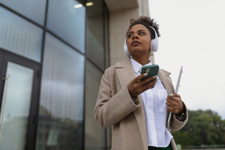 stylish african american female student in headphones using mobile phone and laptop on the background of the glass facade of the building, concept of a strong successful womanの写真素材