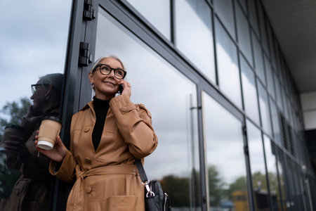Senior woman head in stylish business clothes speaks on a mobile phone against the backdrop of an office buildingの写真素材