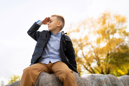 Cheerful mischievous boy sits on a stone and looks into the distance with his hand to his foreheadの写真素材