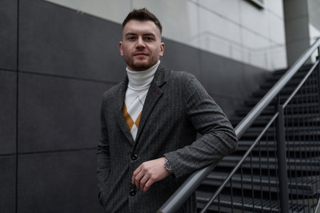 portrait of a strong brutal man in a gray coat against the background of the facade of the building with a staircase leading upの写真素材