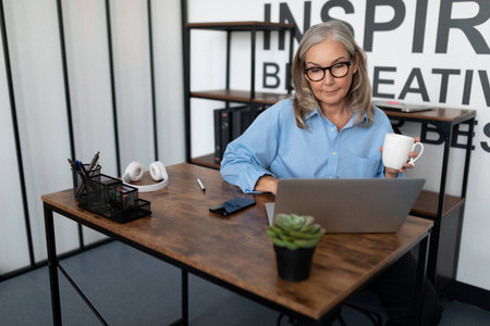 Middle aged older business woman in her fifties working on a laptop online drinking tea in the office, concept of successful business developmentの写真素材