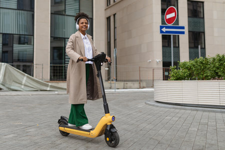 stylish african american woman rides an electric scooter against the backdrop of an office buildingの写真素材