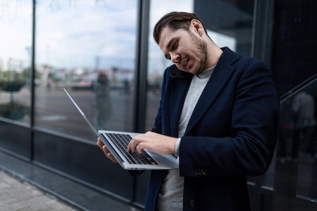 successful male businessman speaks on the phone with his head pressed to his shoulder and typing on a laptopの写真素材