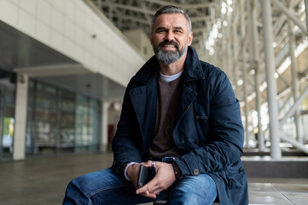 a mature adult male lawyer with gray hair and a beard with a phone in his hands sits in the lobby of the buildingの写真素材