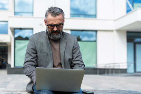 portrait of a mature adult male lawyer with gray hair and beard with a laptop sits on the porch of a buildingの写真素材