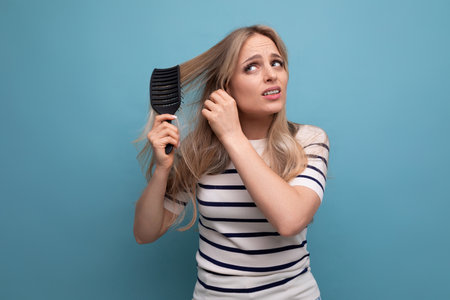 sullen upset blond woman with difficulty combing her hair on a blue backgroundの写真素材
