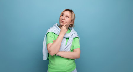 portrait of a blond cute young woman in a casual outfit looks thoughtfully to the side on a studio blue background with copy spaceの写真素材