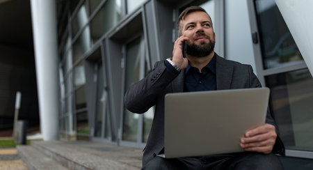 businessman talking on a mobile phone with a laptop on his knees while sitting at the entrance to the office buildingの写真素材