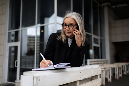 caucasian senior businesswoman taking notes talking on the phone outside the officeの写真素材