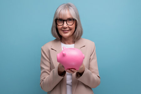 cute old woman in a jacket with a piggy bank of money savings on a bright background with copy spaceの写真素材