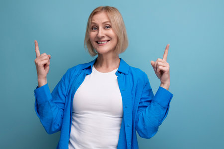 portrait of cheerful blonde 60s woman in casual stylish shirt with crossed arms on studio background
