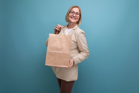 smiling mature woman in jacket holding craft shopping bag on studio background with copy spaceの写真素材