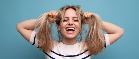 cheerful blond young woman holds her hair in her hands and laughs cutely on a blue backgroundの写真素材