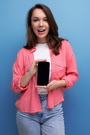 a smiling brunette lady with dark hair below her shoulders in a shirt and jeans demonstrates a smartphone screen forwardの写真素材
