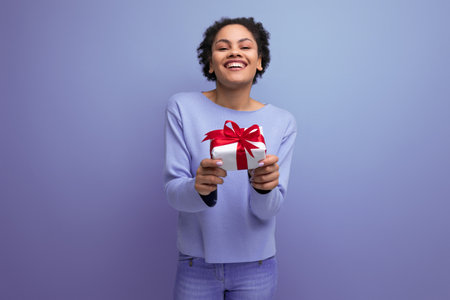 positive young brunette lady with afro curls holds out a gift boxの写真素材