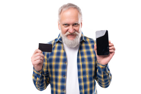 a handsome mature gray-haired man with a beard in a shirt masters online payments with a card and a smartphone on a white background with copy spaceの写真素材