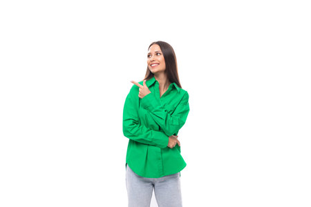 young european female model with well-groomed black hair dressed in a green shirt points her finger to the side on a white background with copy spaceの写真素材