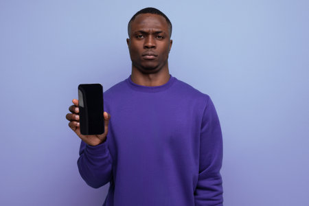 authentic handsome young african man in blue sweater holding smartphone vertically with mockup for advertisement on blue background with copy spaceの写真素材