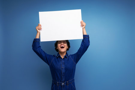 young pretty brunette latin woman with ponytail afro hair dressed in denim clothes reporting the news on a white billboard with advertising spaceの写真素材