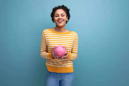 joyful afro brunette 20s latin young woman holding piggy bank of moneyの写真素材