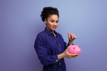young happy brunette latin woman dressed in blue denim overalls accumulated money in a piggy bankの写真素材