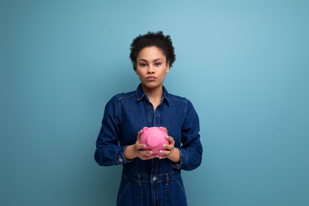 young pretty brunette latin woman with afro hair gathered in a ponytail dressed in denim clothes holding a piggy bank with coins and moneyの写真素材