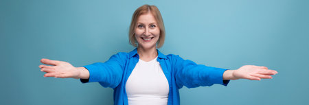 mature woman with blond hair in a casual shirt stretches out her arms hugging on a blue background with copy spaceの写真素材