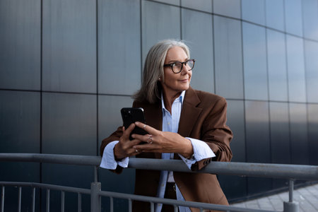 a beautiful gray-haired successful mature business woman in glasses dressed in a respectable way spends a coffee break on the street near the business centerの写真素材