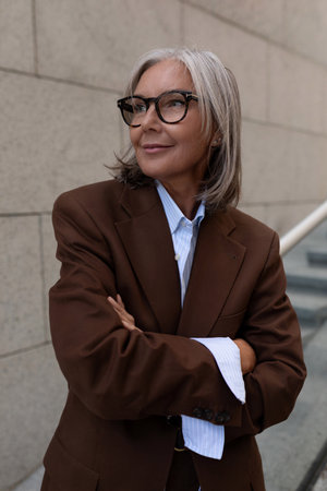 portrait of a slender cute gray-haired entrepreneur woman of mature years in a brown suit on the background of the buildingの写真素材