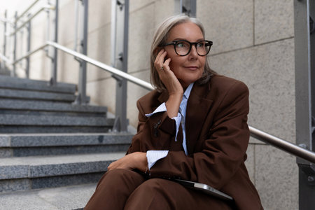 well-groomed slender mature business woman with gray hair is dressed in a brown suit in a city...