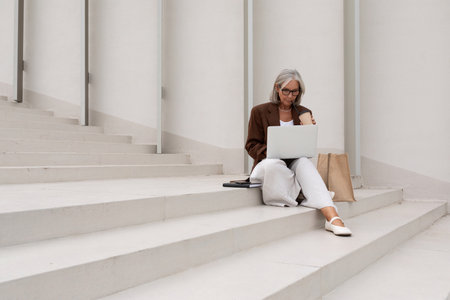 senior business woman with gray hair and glasses dressed in a jacket works remotely with a laptop sitting on the stairsの写真素材